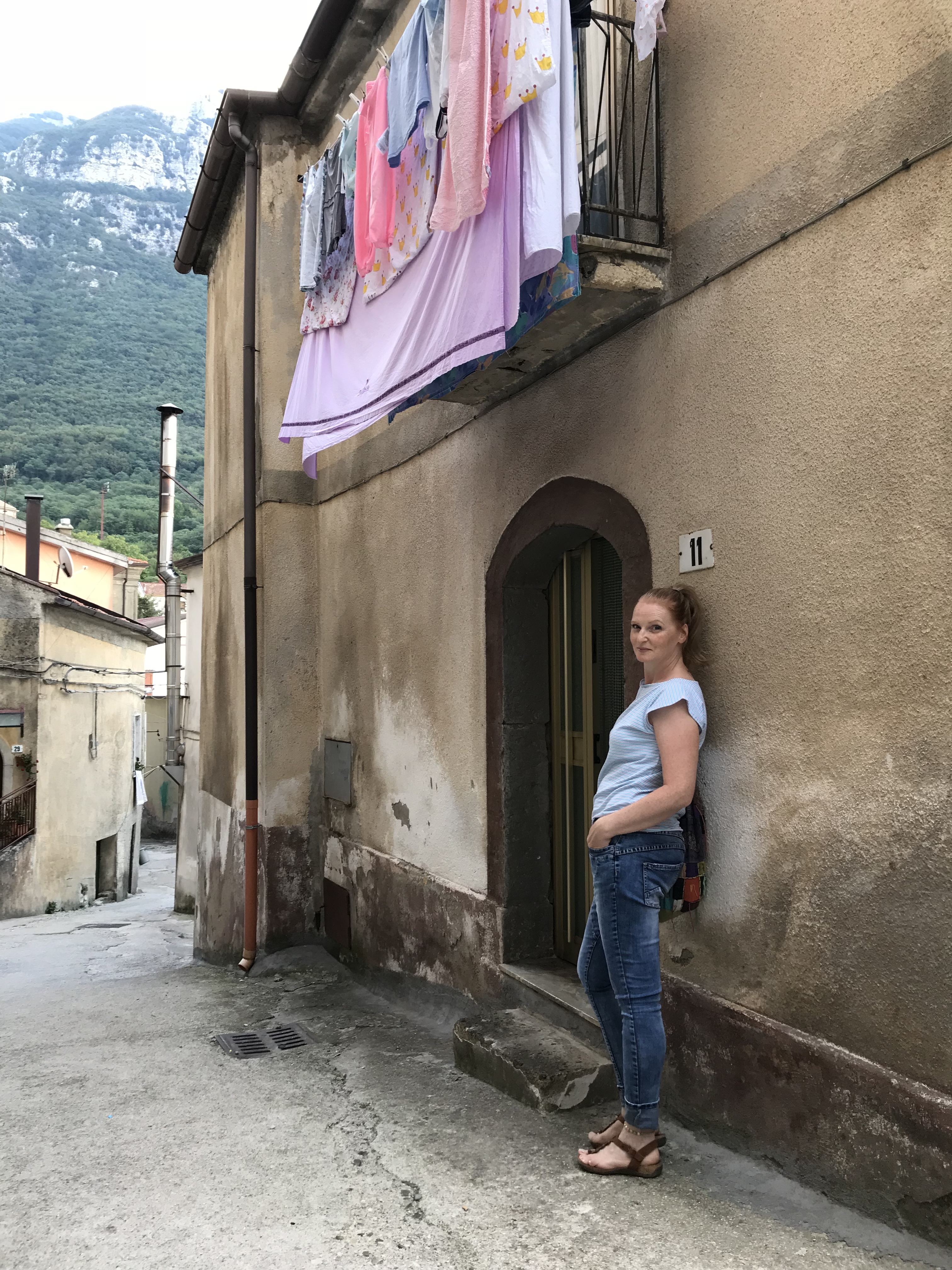 Giovanna in einer italienischen Dorfgasse vor einem alten Haus und den Bergen